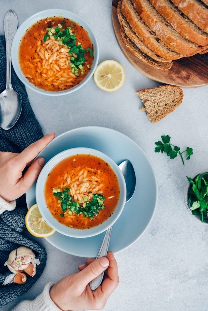 Turkish Tomato Orzo Soup Recipe packs a ton of flavor with fewer than 10 ingredients in just 20 minutes. The combination of tomatoes, lemon, parsley and orzo makes the easiest pasta soup ever. #orzosoup #wintersoup #souprecipe #turkishsoup Woman eating Turkish tomato orzo soup garnished with parsley served in light blue bowls on a grey background.