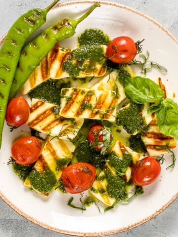 Grilled halloumi cheese, tomatoes and green peppers drizzled with a herbed oil sauce on a white plate.