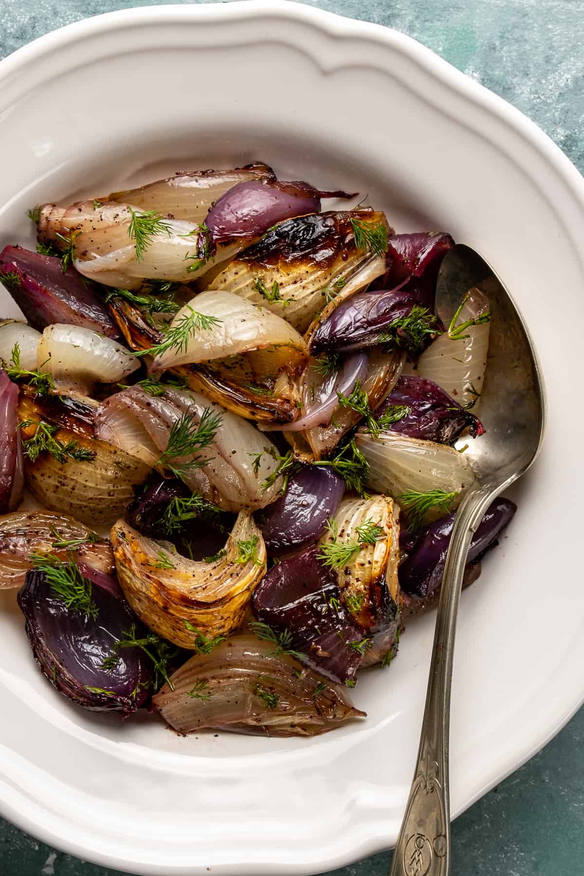 Red onions and yellow onions are roasted and combined with fresh dill and pomegranate molasses in a white bowl and a spoon inside it.