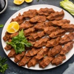 Turkish cigkofte with fine bulgur and spices on a white plate with parsley and lemon wedges and pomegranate molasses and lettuce leaves on the side.