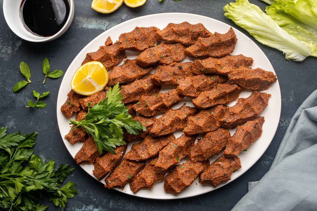 Turkish cigkofte with fine bulgur and spices on a white plate with parsley and lemon wedges and pomegranate molasses and lettuce leaves on the side.