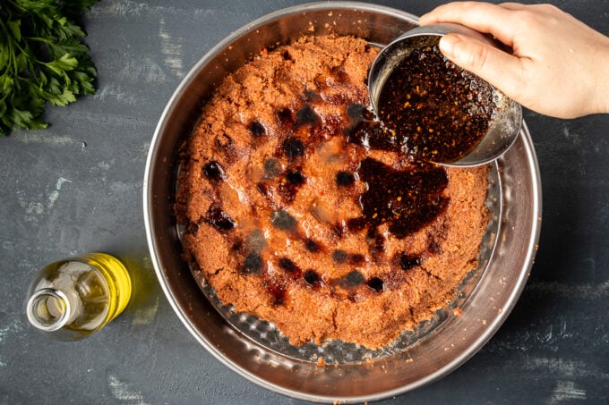 A hand pouring a mixture of olive oil and isot pepper on a bulgur mixture in a traditional pan.
