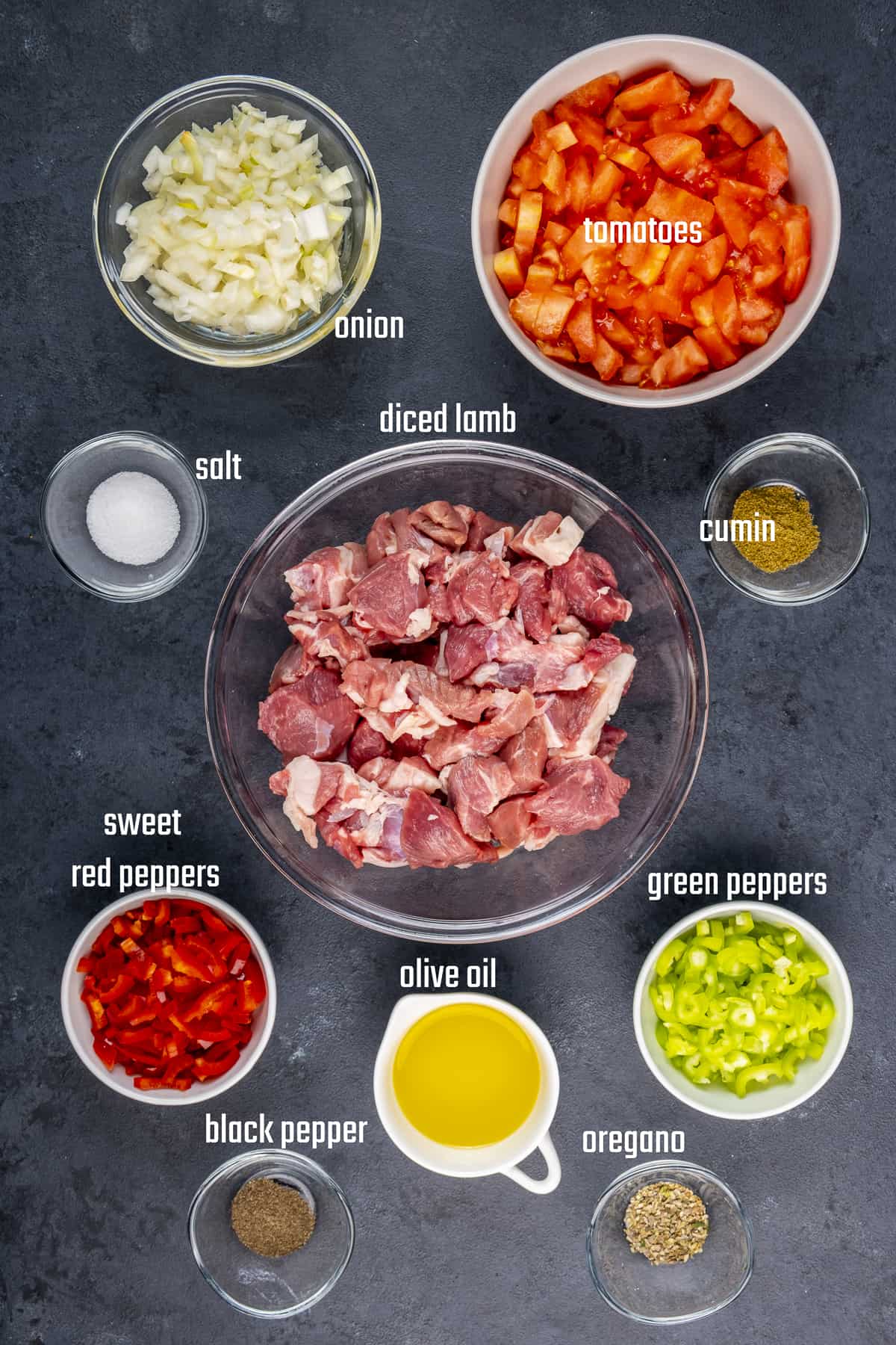 Diced lamb, chopped tomatoes, peppers, onion, olive oil, salt, pepper, cumin and oregano on a black backdrop.