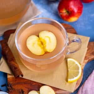 Apple tea in a glass mug, a slice of apple inside it, a lemon slice and cinnamon stick on the side.