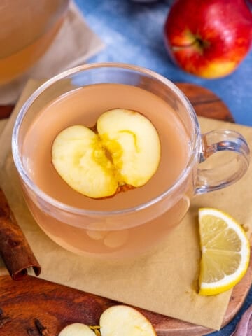 Apple tea in a glass mug, a slice of apple inside it, a lemon slice and cinnamon stick on the side.