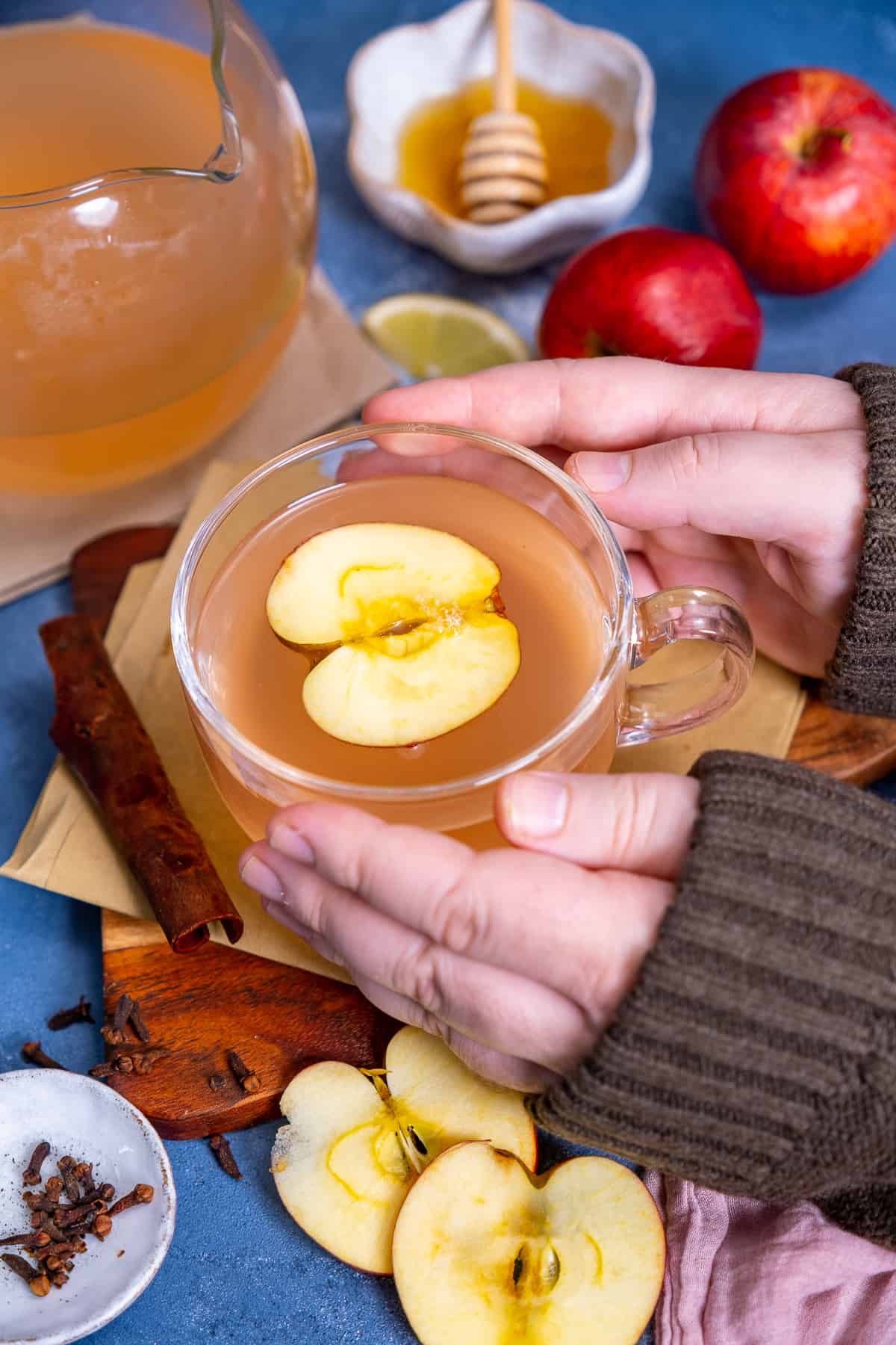 Hands holding a cup of warm apple tea, apples and honey on the side.