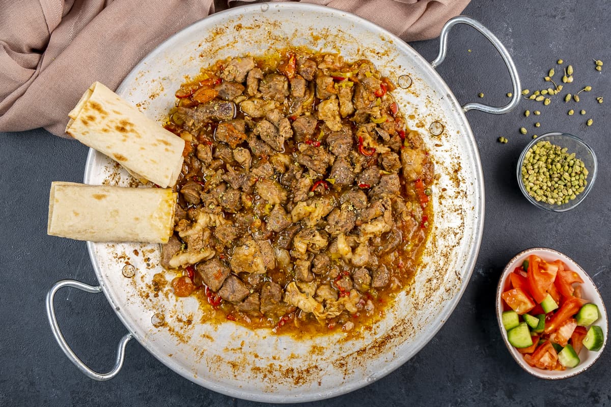 Sac kavurma in a traditional pan accompanied by tomato and cucumber salad and oregano in a small bowl.