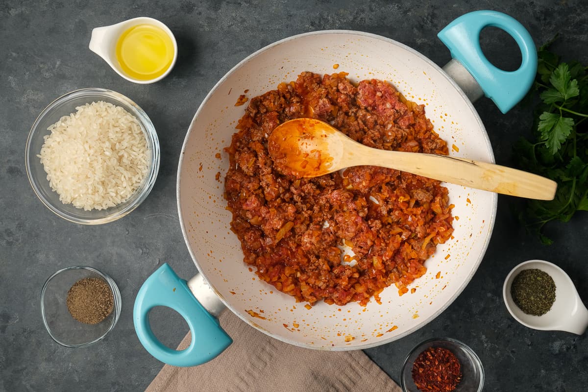 Ground beef and onions cooking with tomato paste in a white pan and a wooden spoon inside it.
