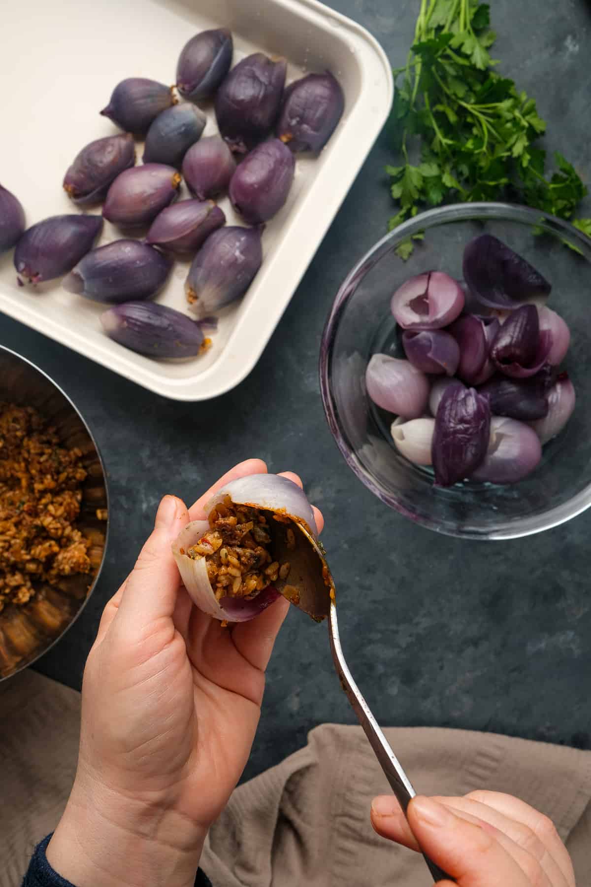 Hands stuffing an onion with ground beef and rice filling with a spoon more stuffed onions in a pan and onion layers in a bowl on the side.