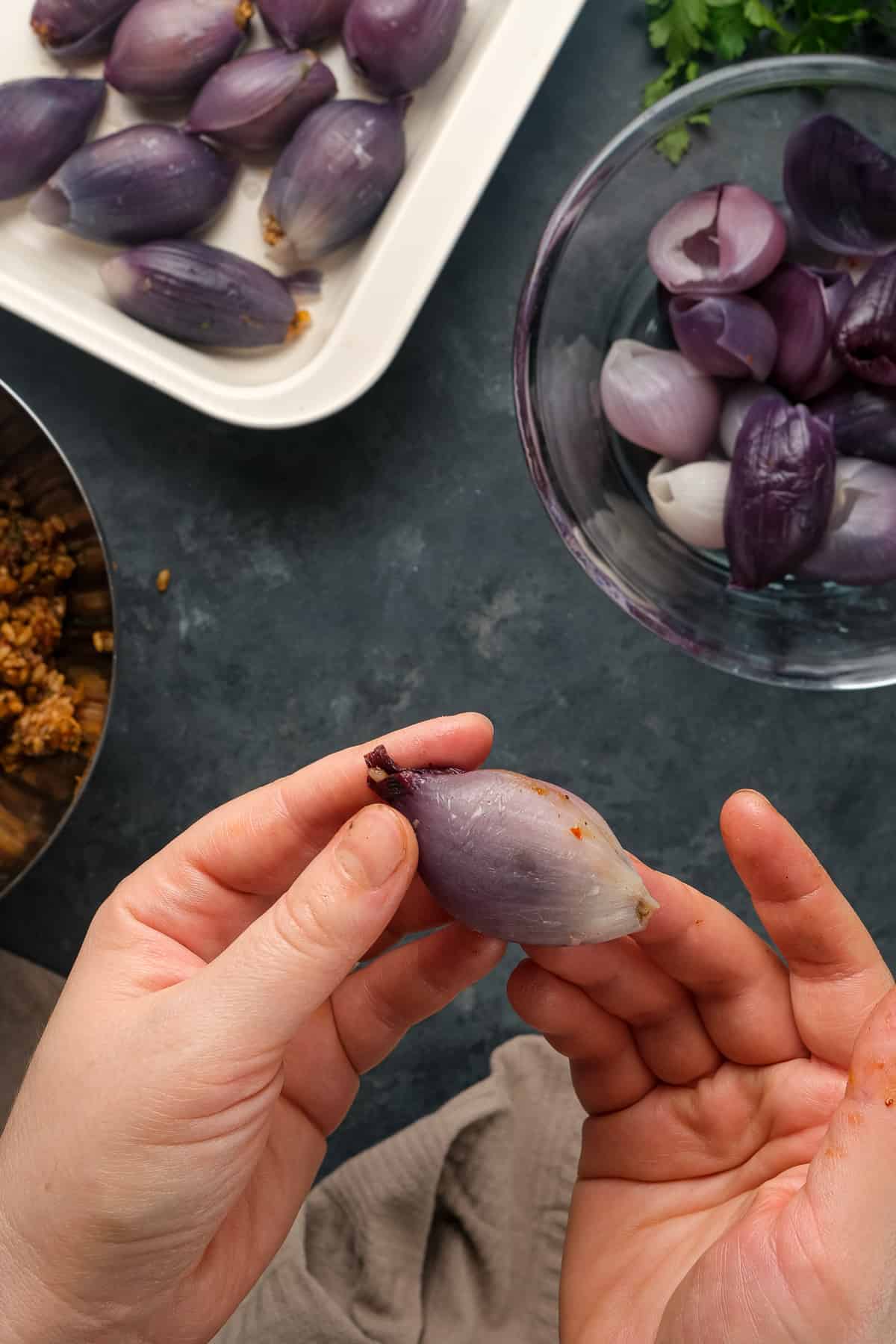 Hands holding a stuffed purple onion and more stuffed onions in a pan, filling in a bowl and cooked onions in another bowl on the side.