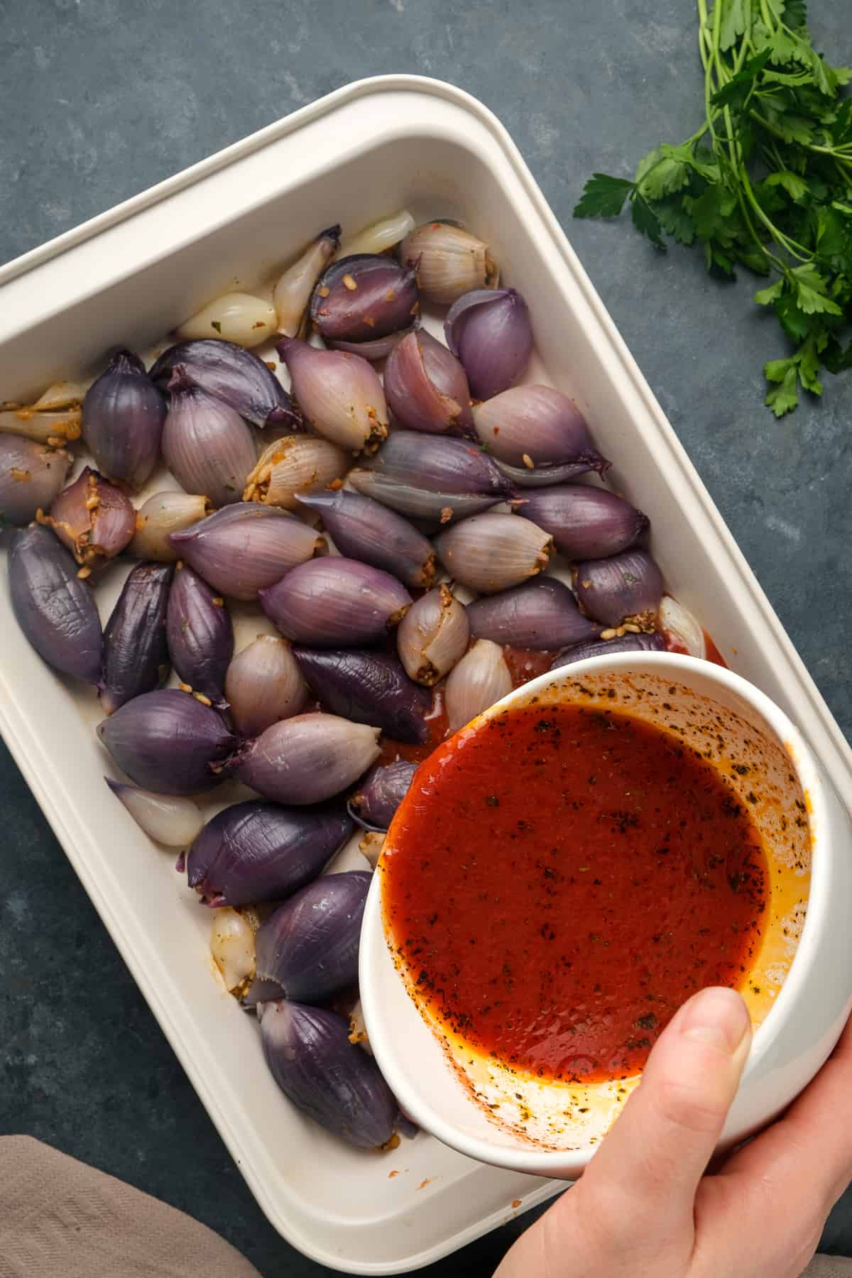 A hand pouring tomato sauce over stuffed purple onions in a baking pan.