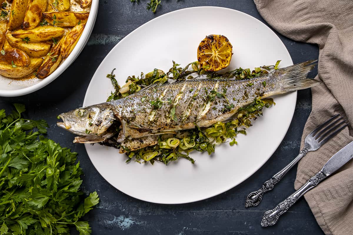 Oven baked sea bass served on a white plate with roasted leeks and baked potatoes in another pan on the side.