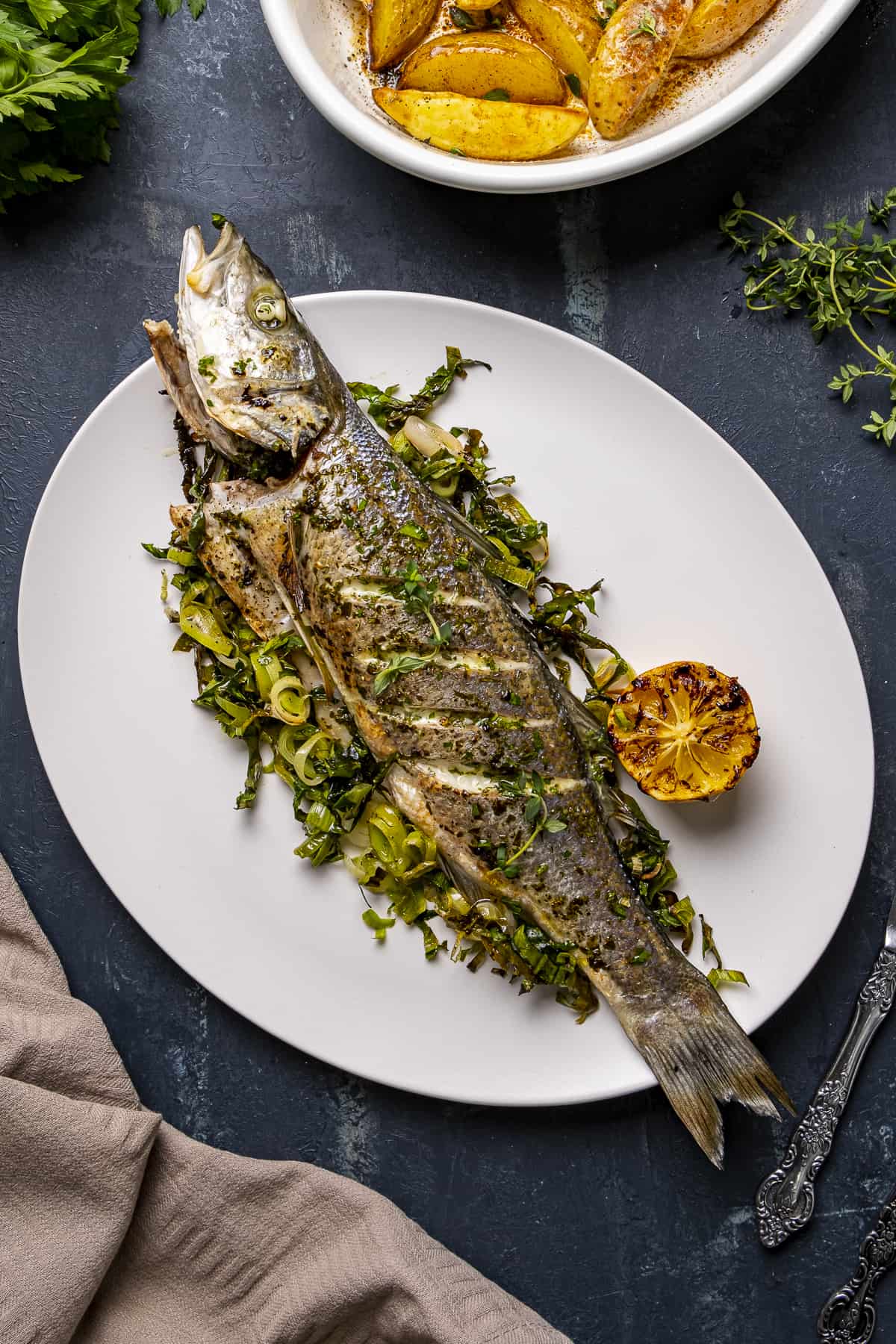 Baked sea bass served on a white plate with some roasted green vegetables and lemon and roasted potatoes in another pan on the side.