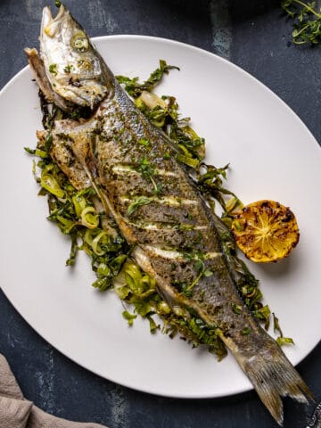 Whole sea bass baked in oven served on roasted green vegetables on a white plate.