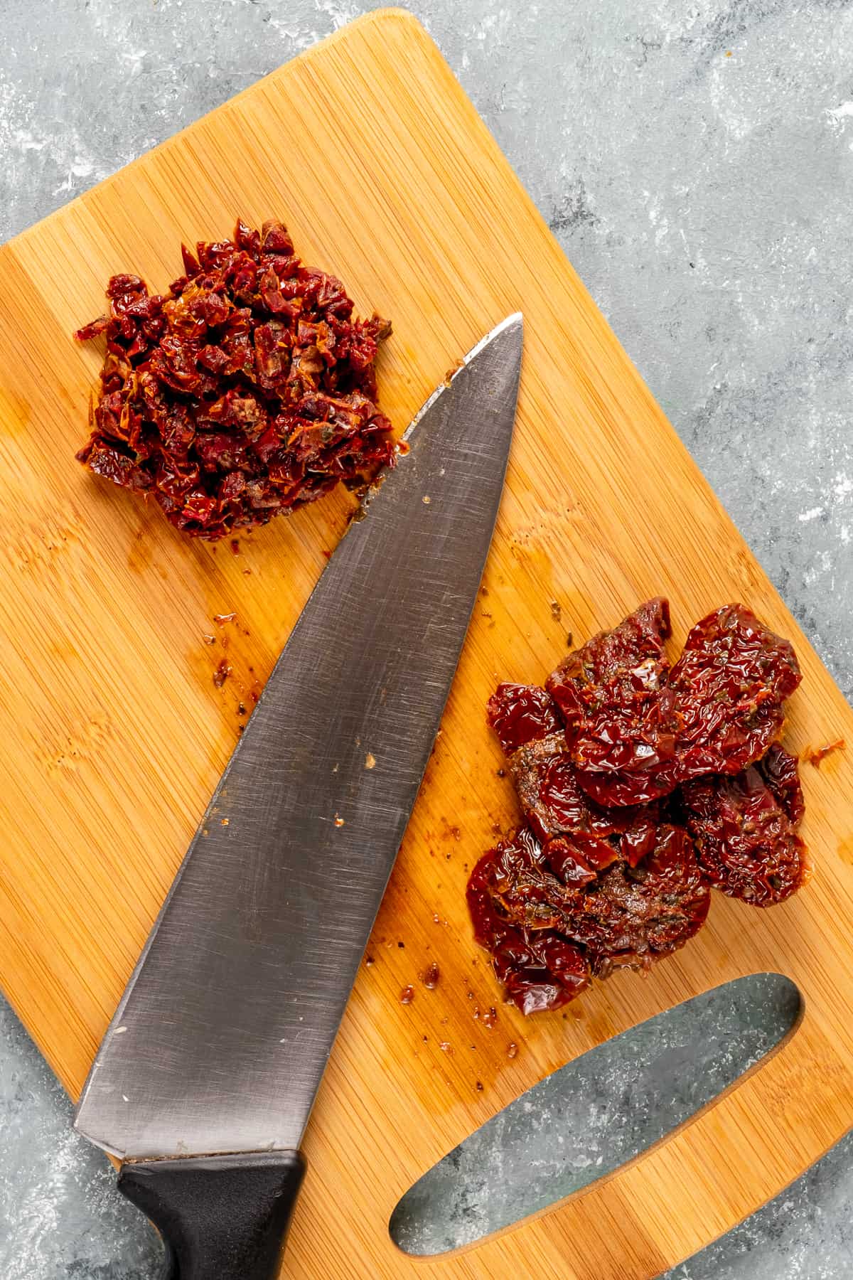 Sun-dried tomatoes whole and finely chopped on a wooden board and a knife in between them.