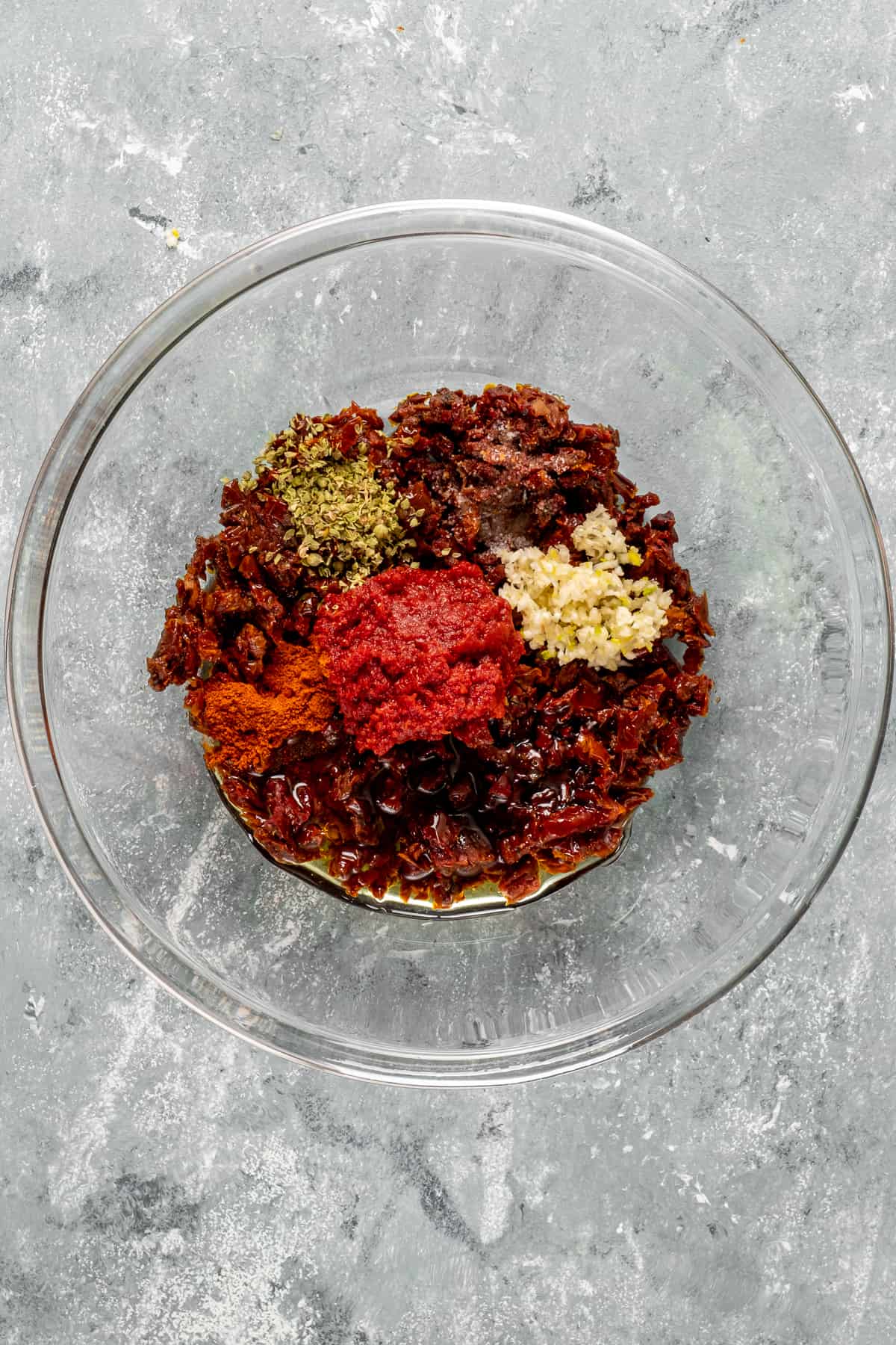 Spices, chopped sun-dried tomatoes, minced garlic and tomato paste in a glass mixing bowl.