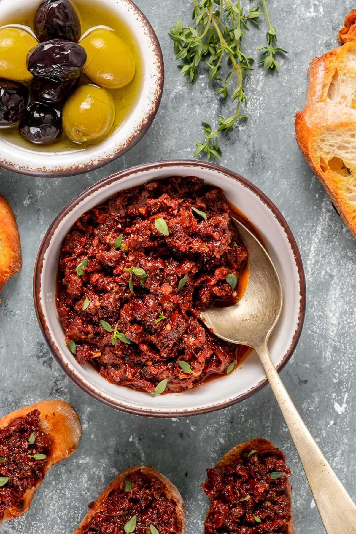 Sun-dried tomato sauce in a white bowl with a spoon inside it and black and green olives, fresh thyme leaves and toasted bread accompany.