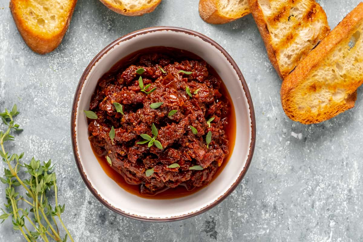 Spicy dip with sun-dried tomatoes in a white bowl, toasted bread slices and fresh thyme on the side.