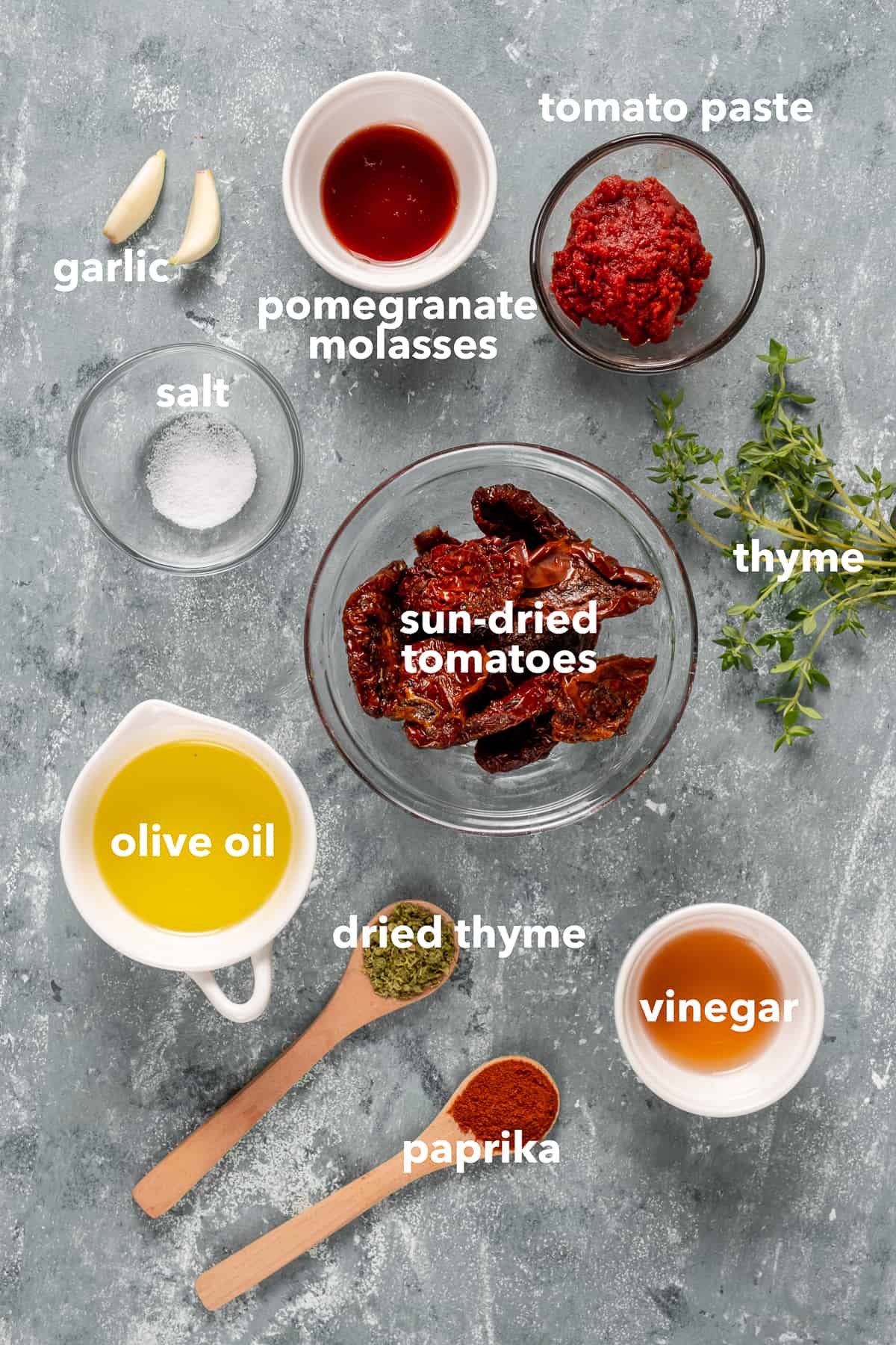 Sun dried tomatoes, tomato paste, pomegranate molasses, olive oil, vinegar, spices, garlic and fresh thyme are on a grey background.