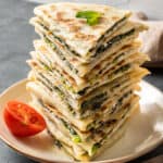 Spinach and cheese filled gozleme served in a stack on a white plate and a half of a cherry tomato on the side.