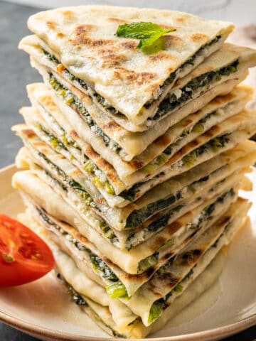 Spinach and cheese filled gozleme served in a stack on a white plate and a half of a cherry tomato on the side.
