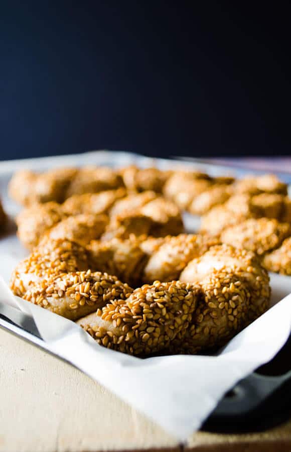 Simit dough coated with molasses and sesame seeds on a baking paper lined baking sheet from front view and more simits behind it.