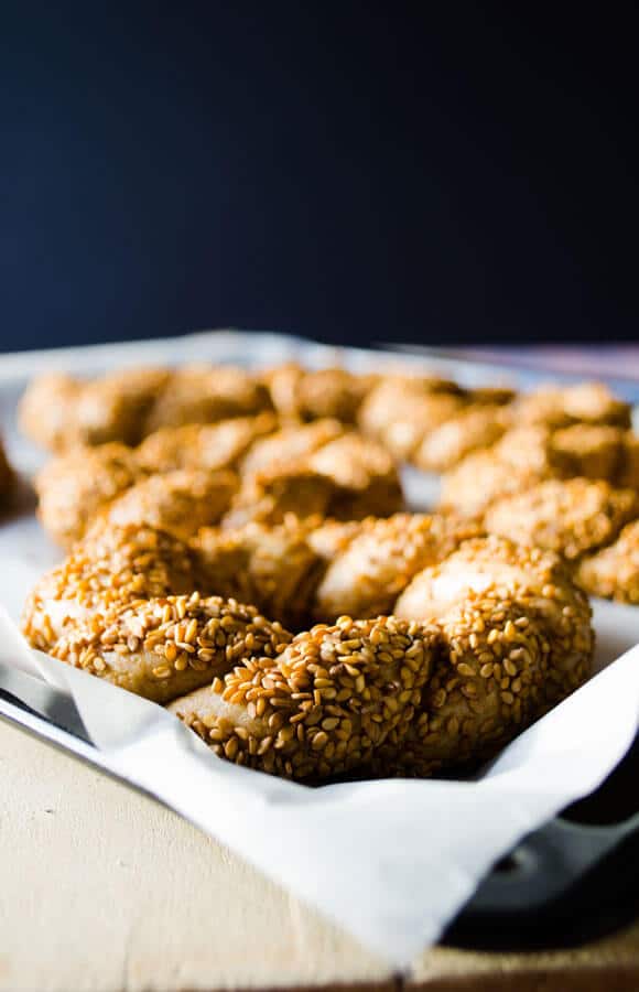 Simit dough coated with molasses and sesame seeds on a baking paper lined baking sheet from front view and more simits behind it. 