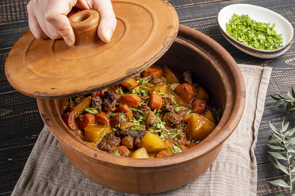 A hand holding the lid of the clay pot that has beef stew inside.