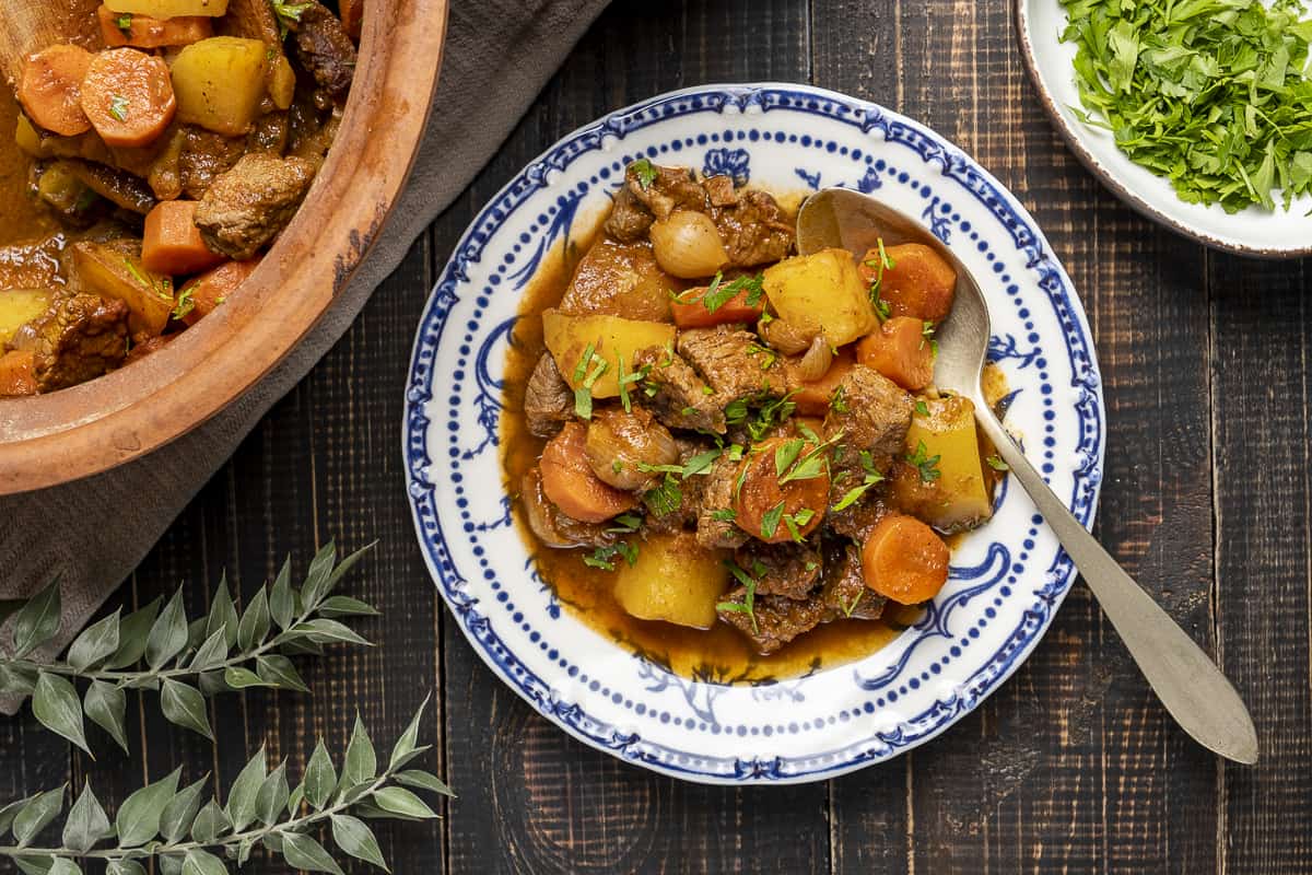 Beef stew with potatoes and carrots served on a white and blue plate and the clay pot is on the side.