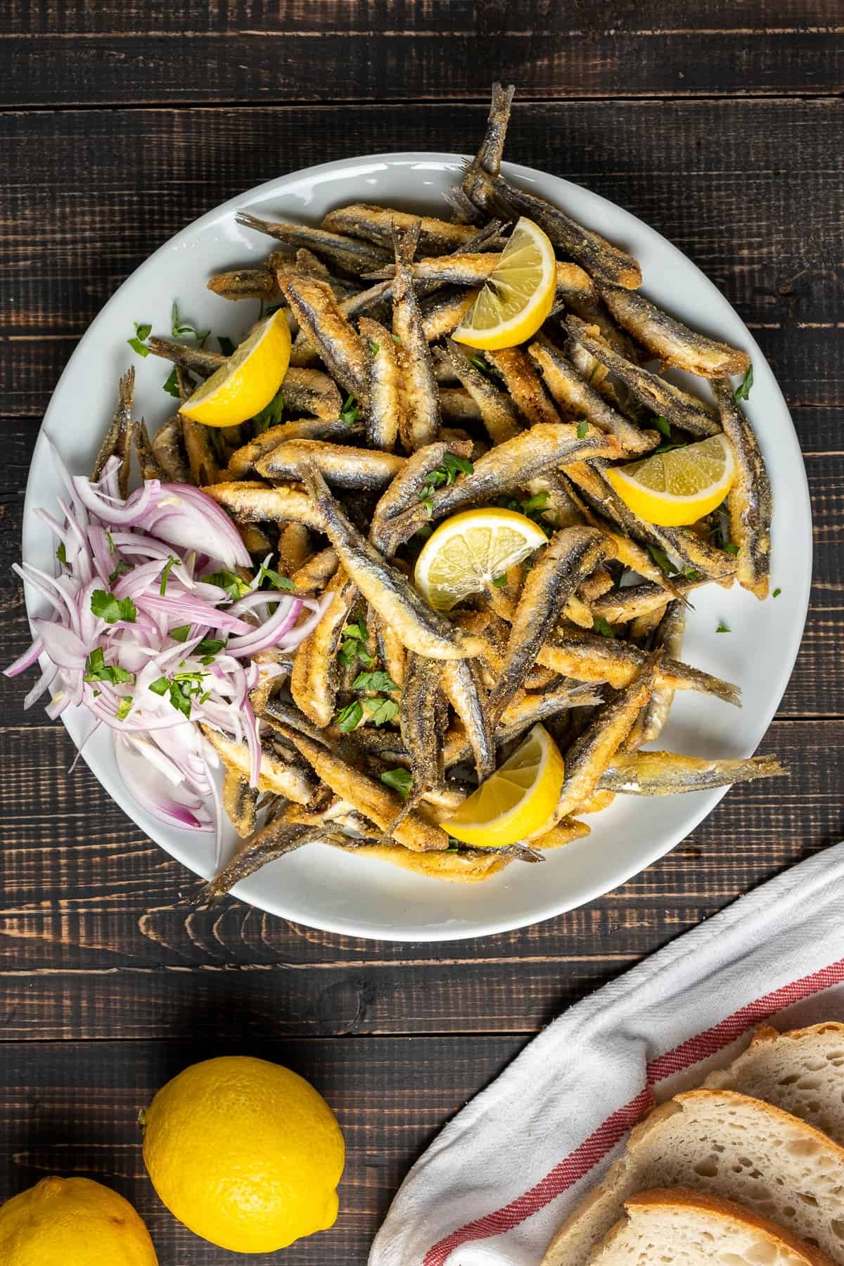 Fried anchovies with lemon wedges and onion slices on a plate and bread slices on the side.