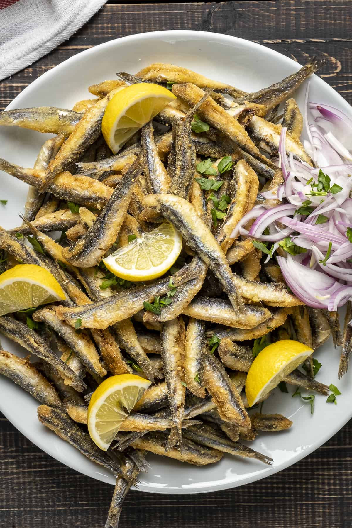 Fried anchovy fish on a plate with lemon wedges and sliced onions.
