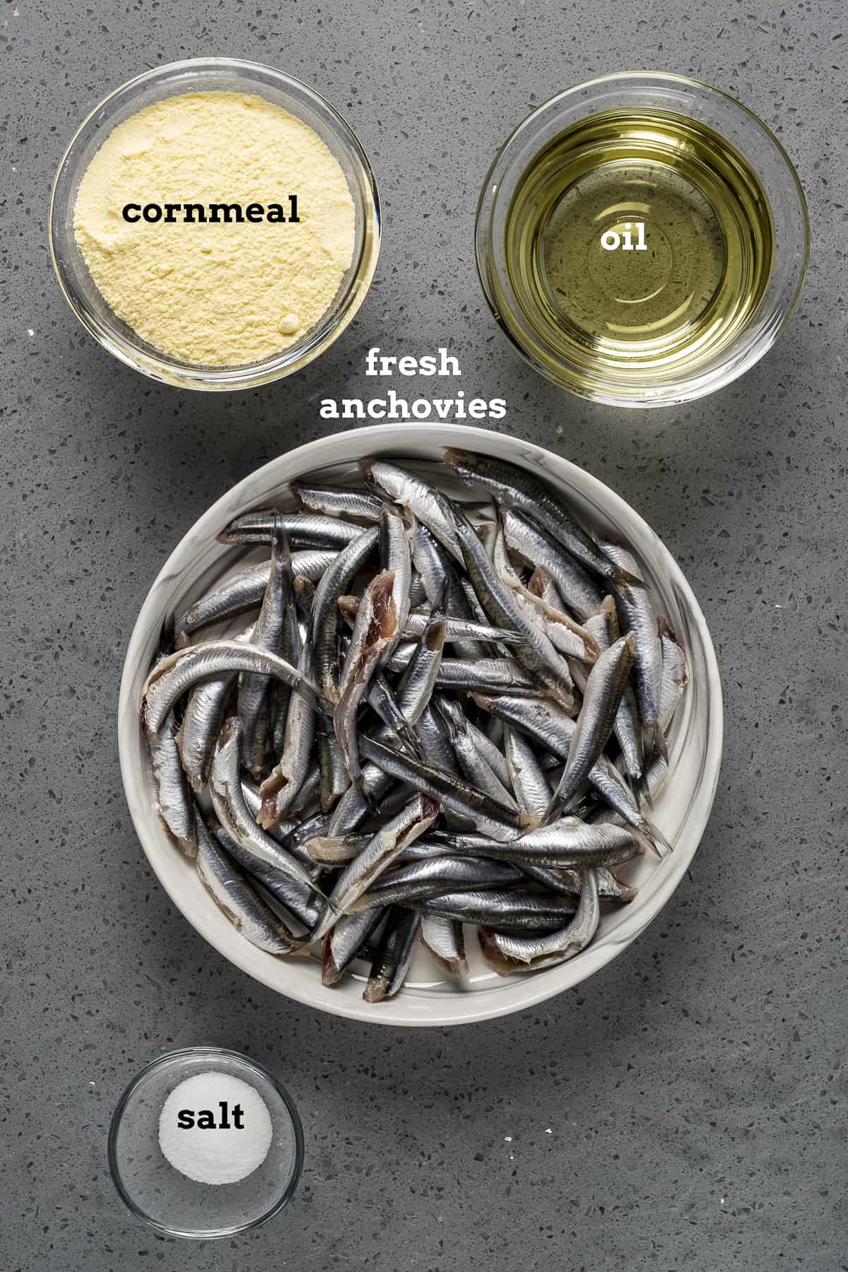 Fresh anchovies, cornmeal, oil and salt on a grey background.