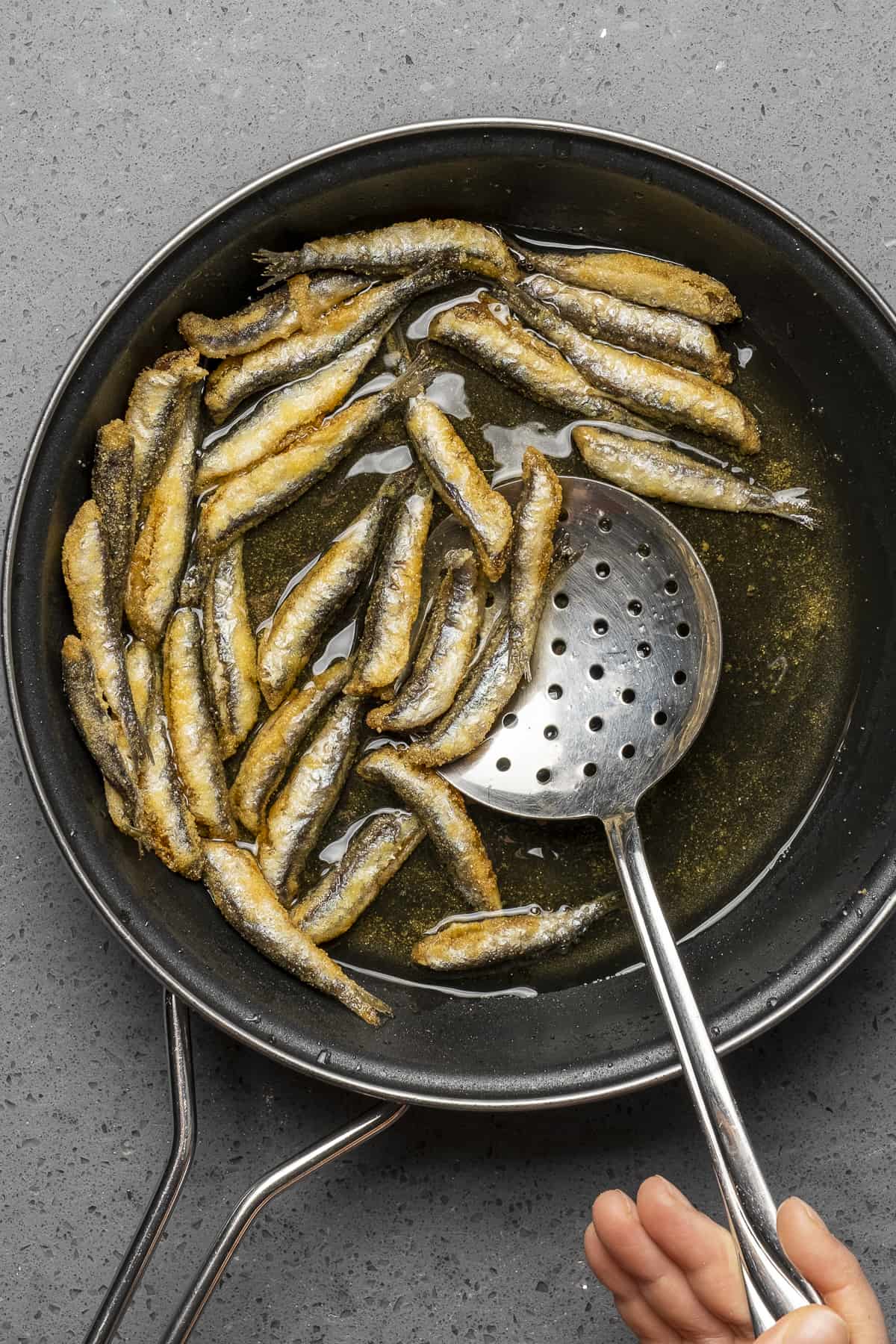 Hand removing fried anchovies from a frying pan with a slotted spoon.