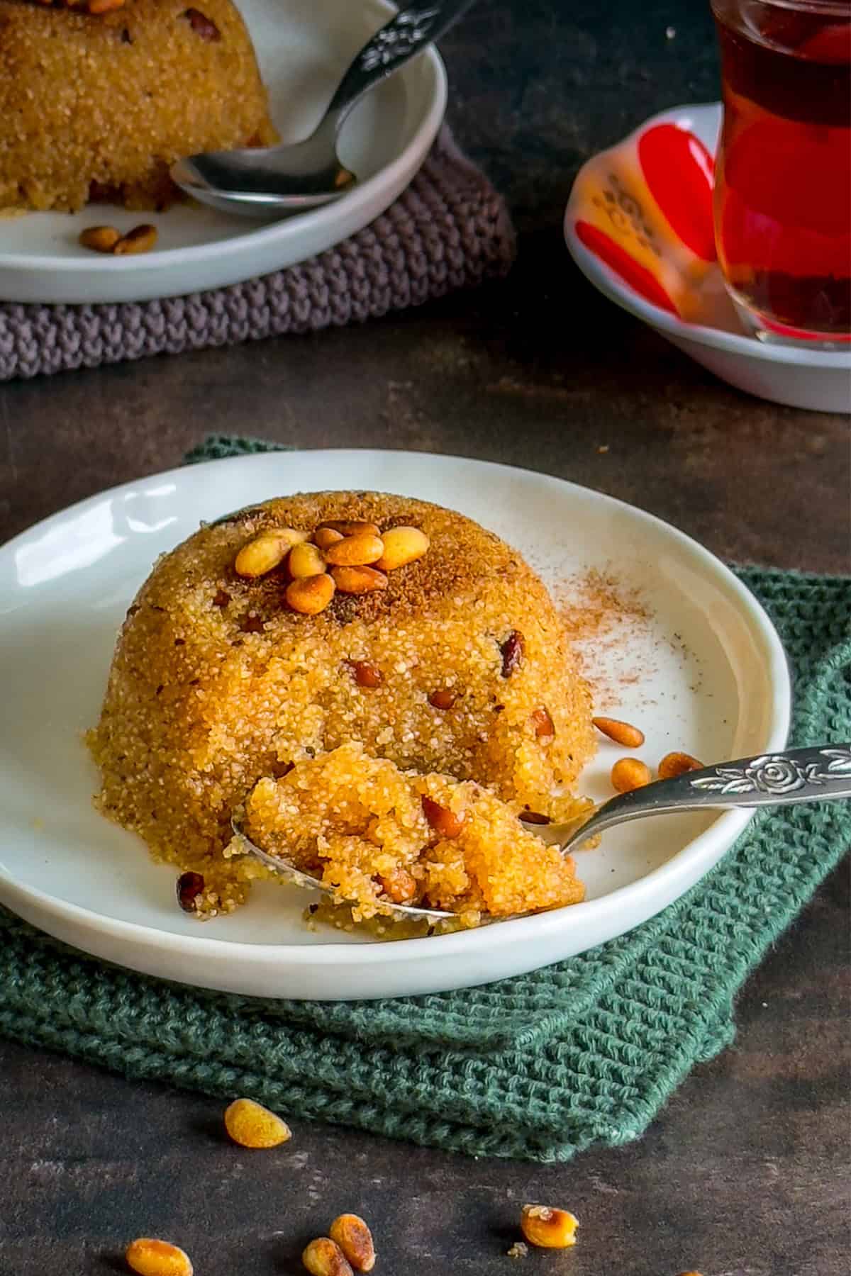Turkish semolina halva (irmik helvasi) with pine nuts on a white plate, served with a spoon and a glass of tea.