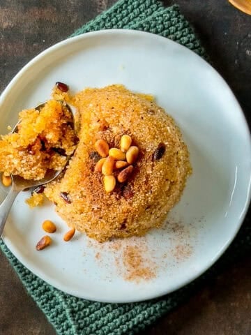 A dome-shaped serving of Turkish semolina halva (irmik helvasi) topped with pine nuts and cinnamon on a white plate, with a spoonful taken out.