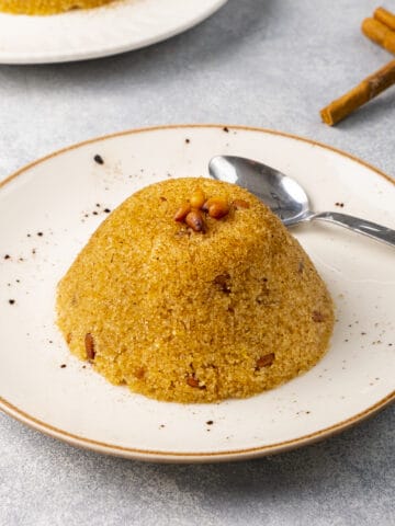 Semolina halva topped with toasted pine nuts served on a white plate with a spoon on the side.