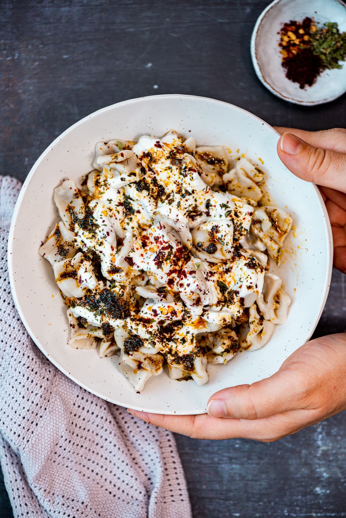 Hands holding a bowl of manti with yogurt and spicy butter sauces on a dark background.