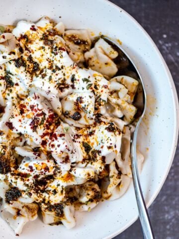 Turkish manti with a yogurt sauce topped with a spicy butter sauce in a white bowl and a spoon inside it.