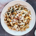 Manti dumplings with a generous amount of yogurt,melted butter and spices in a white bowl on a dark background. Spices in a tiny white bowl on the side.