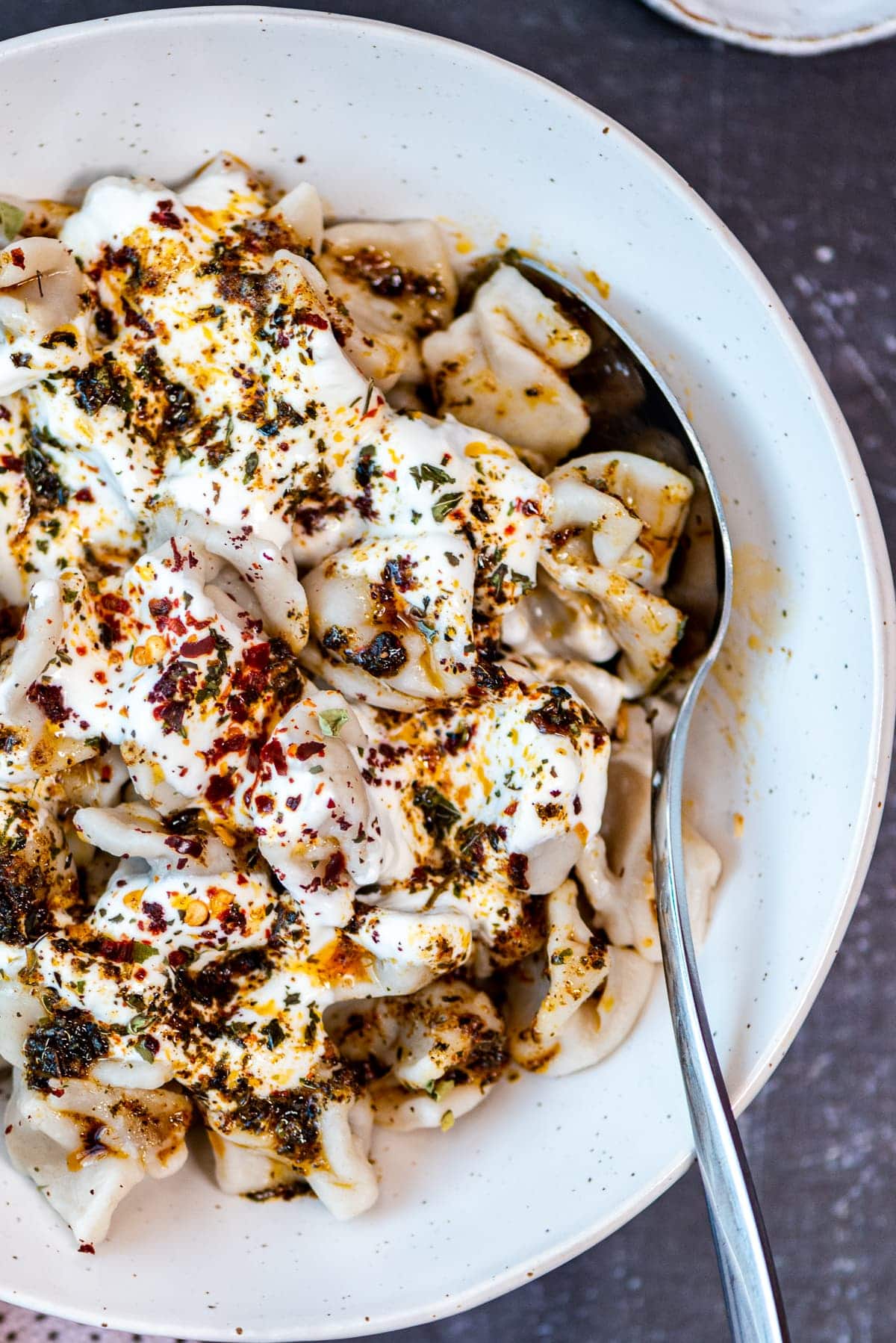 Turkish manti with a yogurt sauce topped with a spicy butter sauce in a white bowl and a spoon inside it.