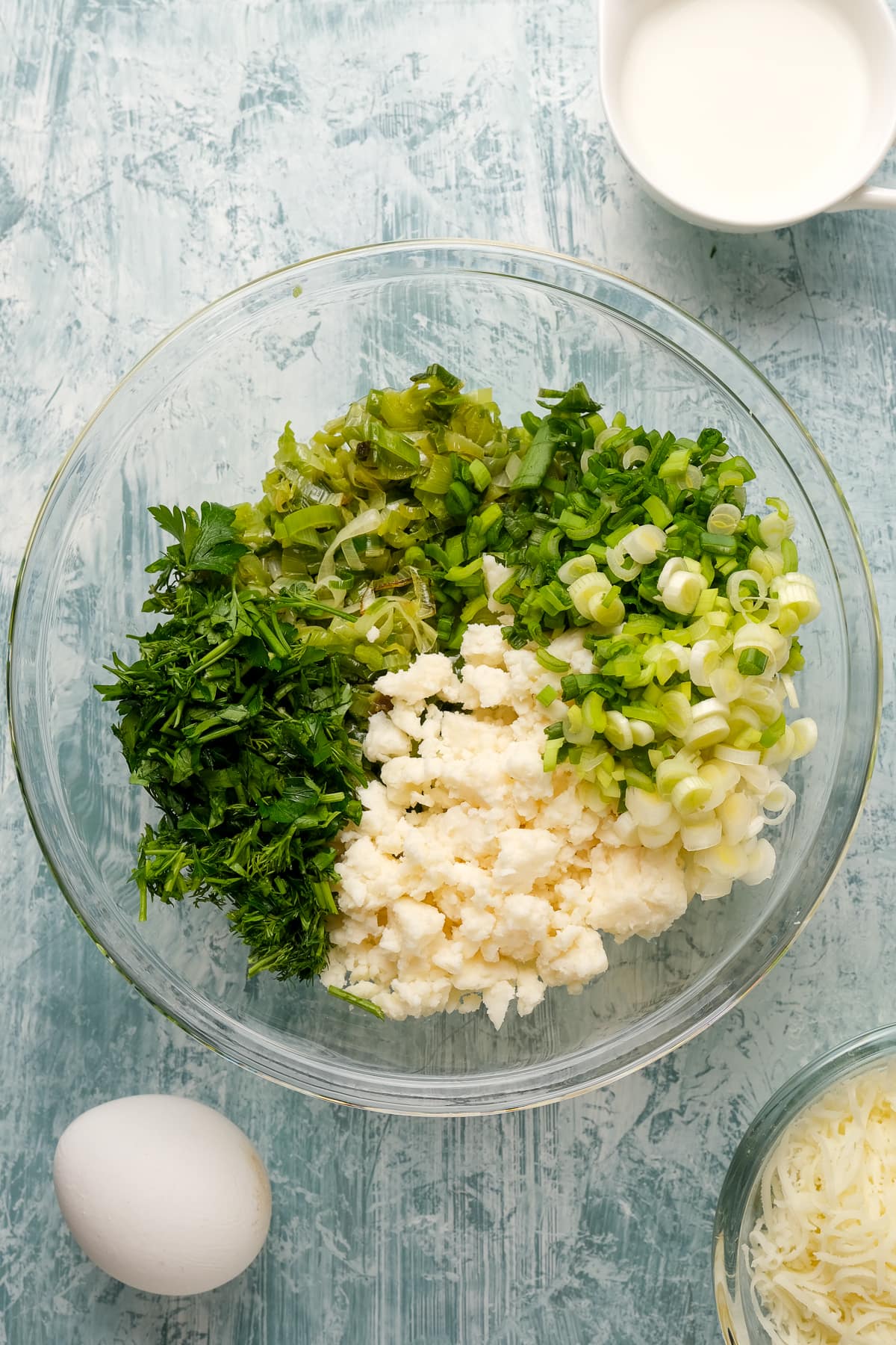 Cooked leeks, green onions, herbs and cheese in a glass bowl on a light background.