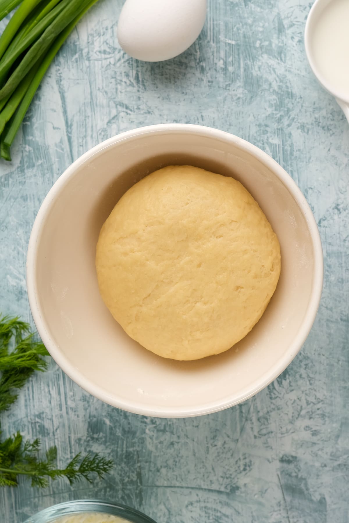 Crust dough ready in a white bowl, an egg, green onions and herbs on the side on a light background