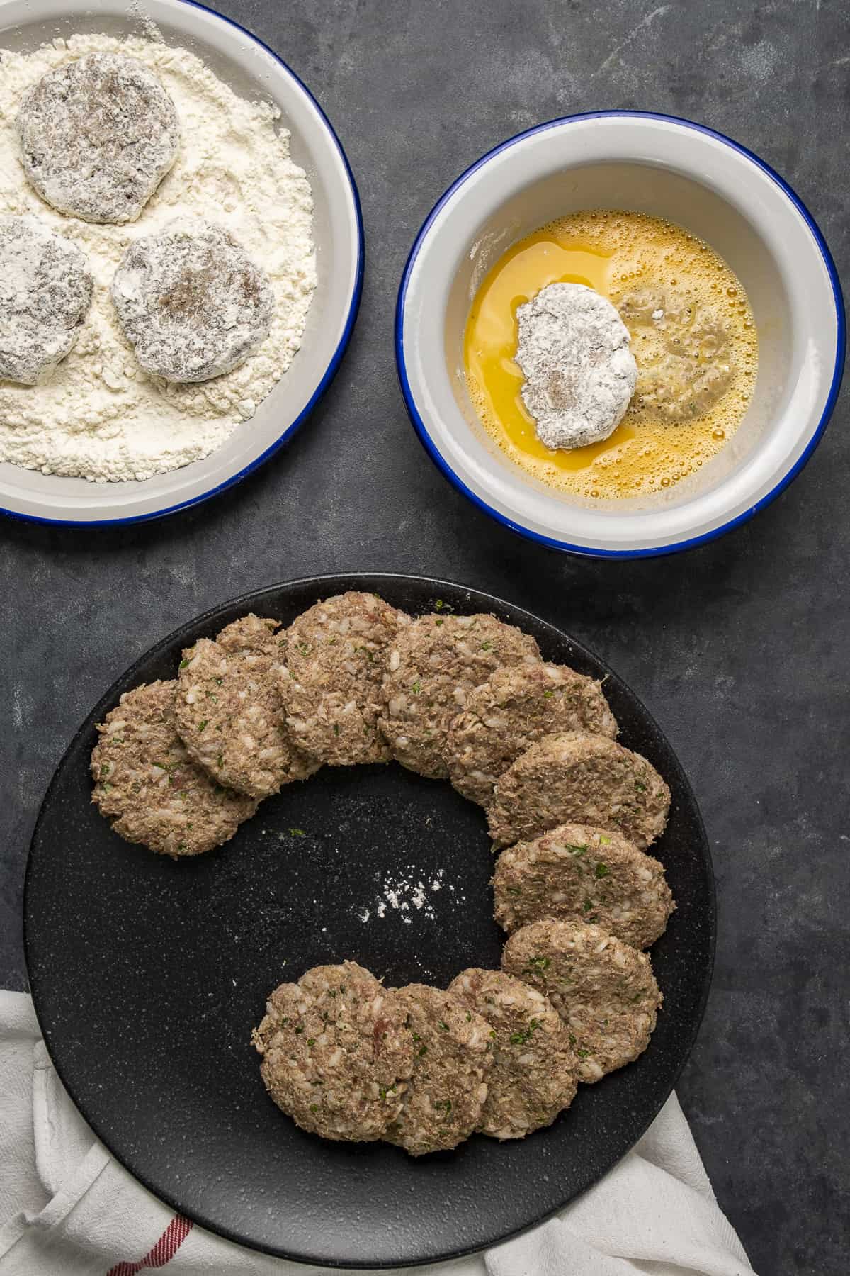 Uncooked meatballs on a black plate, a few meatballs coated with flour amd two meatballs coated with egg in another bowl.