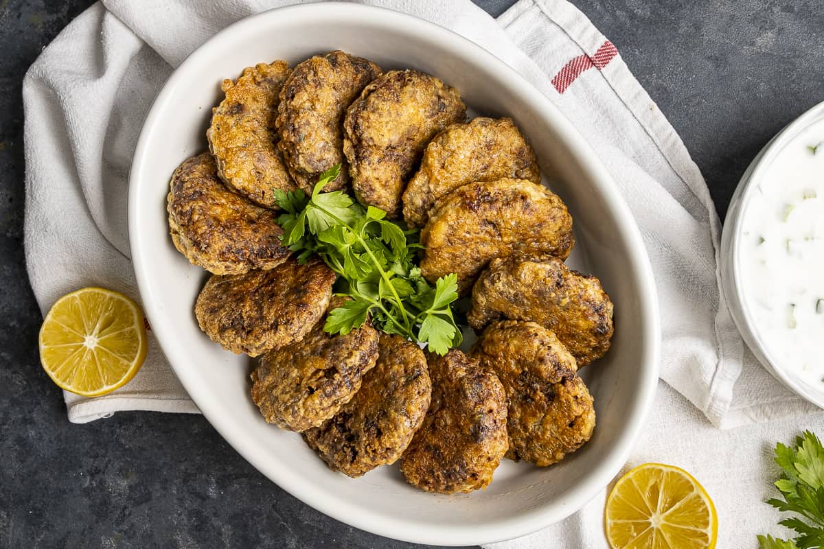 Kadinbudu kofte in a white oval dish, lemon halves on the ground and a bowl of yogurt dip on the side.