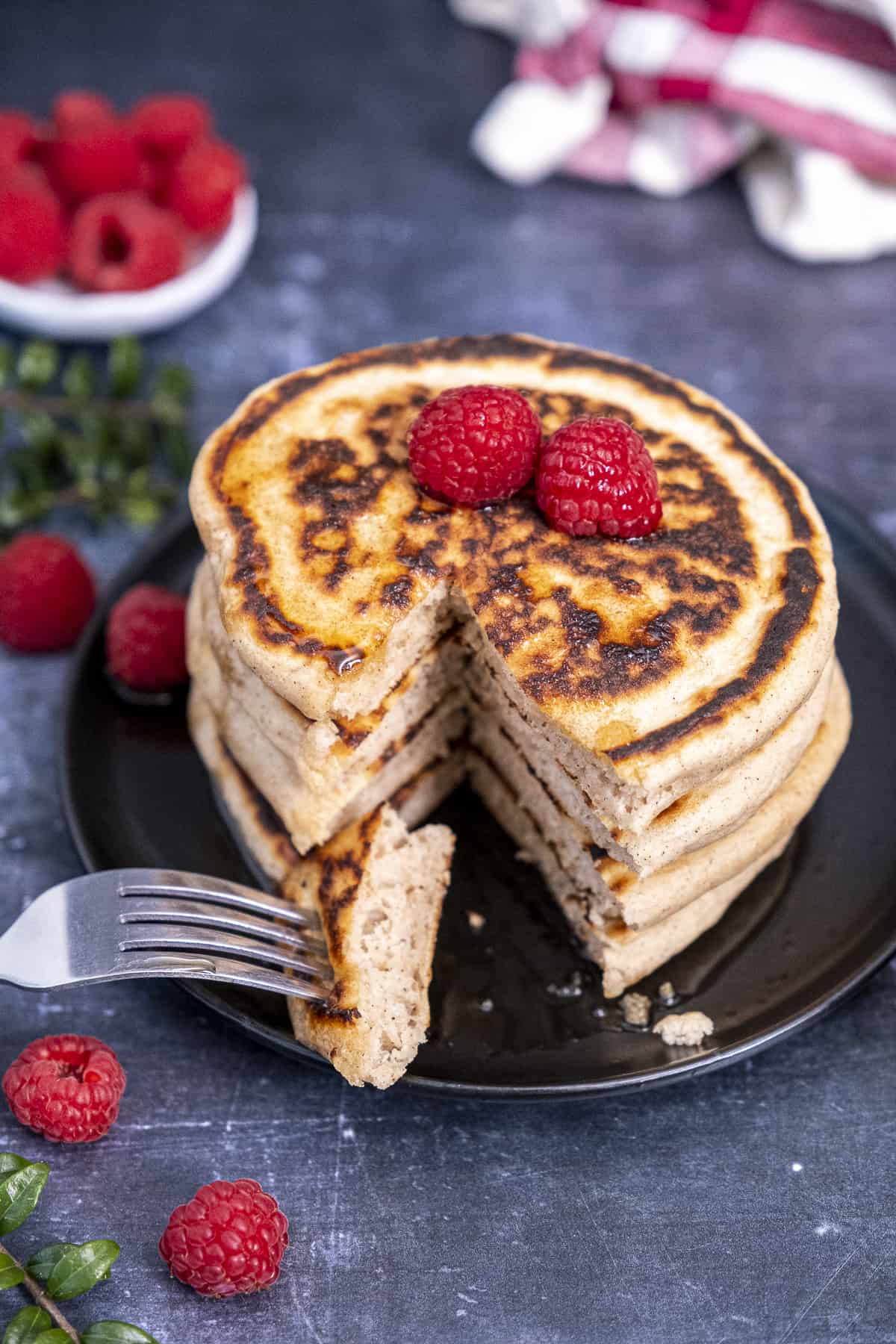 A stack of pancakes sliced and a piece of pancake on a fork on a black plate, raspberries on the top.