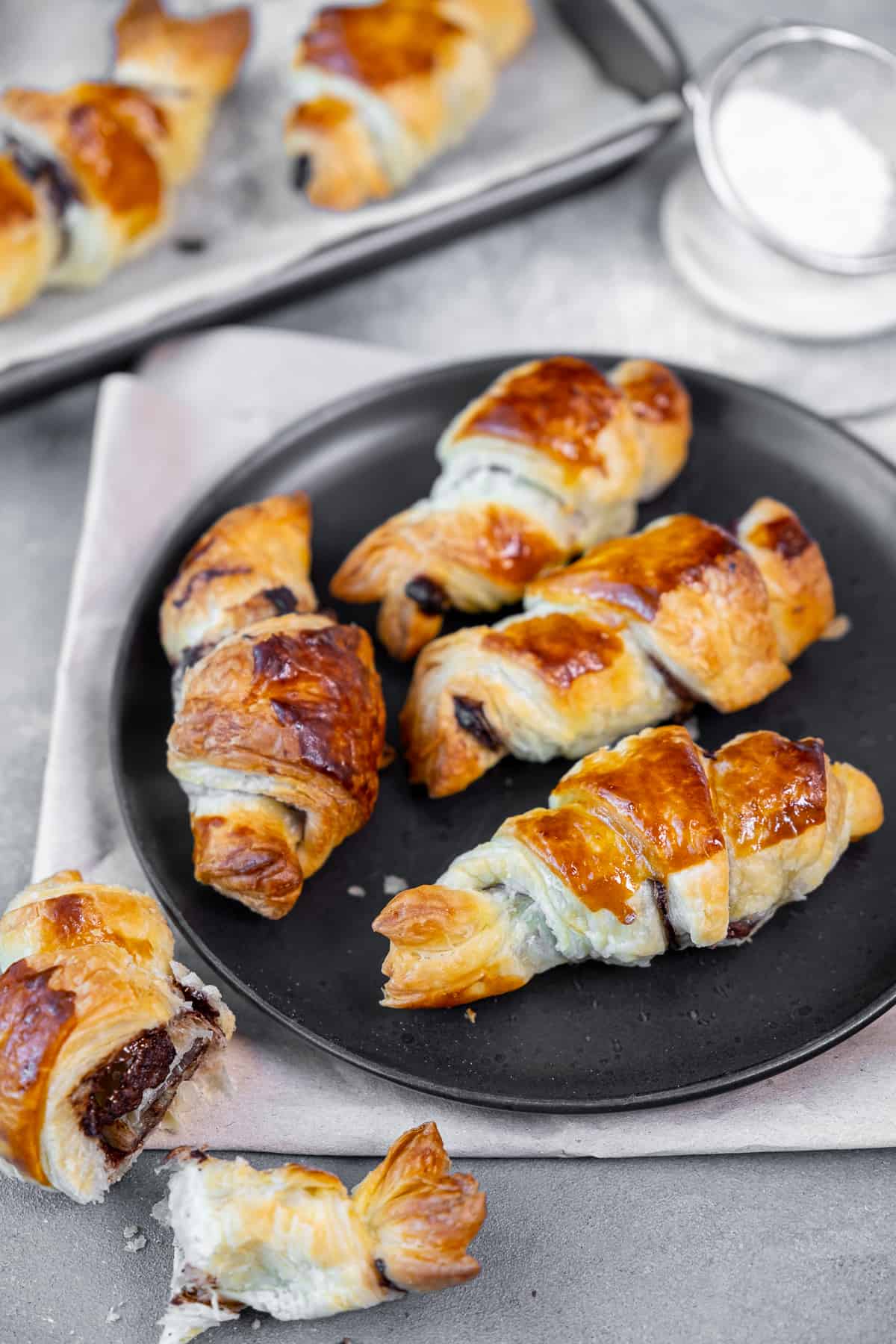 Chocolate filled puff pastries on a black plate, powdered sugar and more pastries behind.