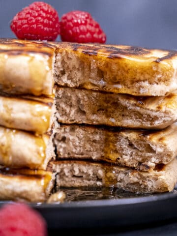 Sliced fluffy pancakes drizzled with maple syrup and topped with raspberries on a black plate.