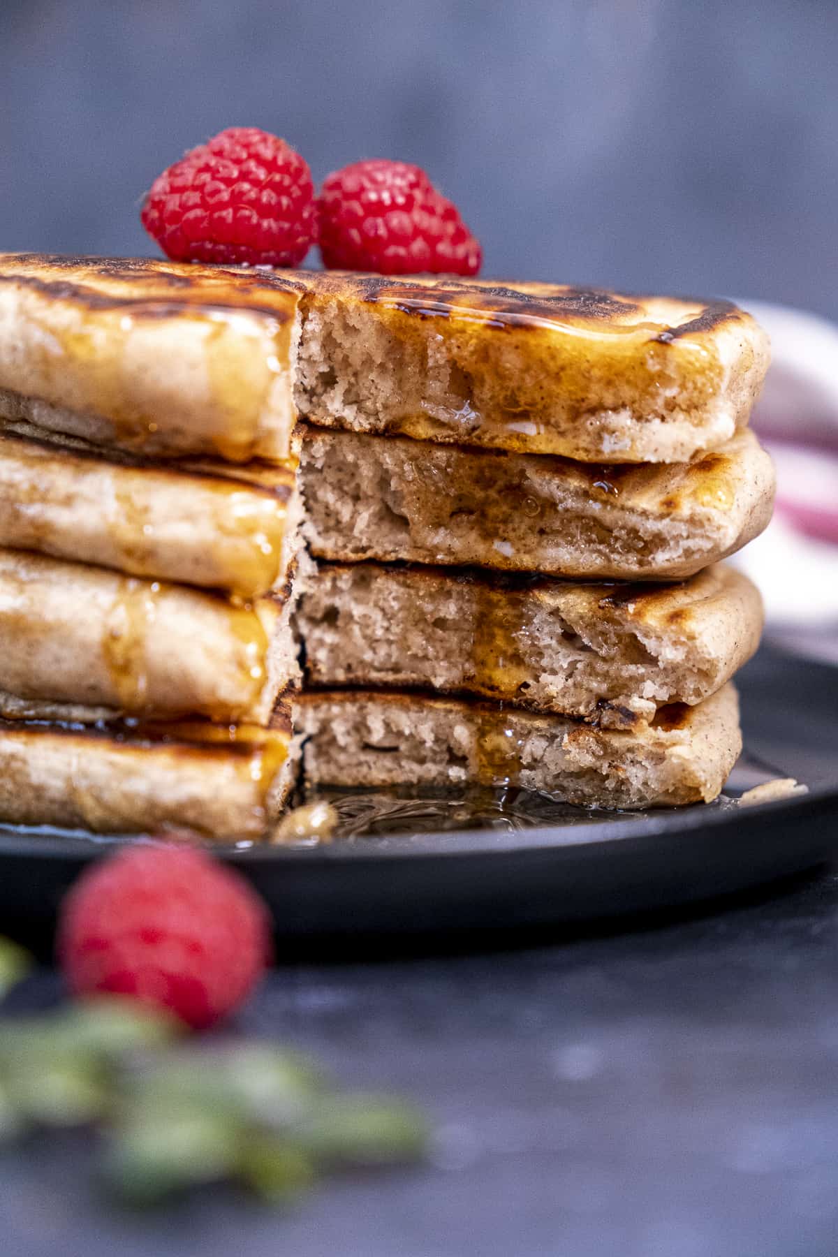 Fluffy pancakes sliced, drizzled with maple syrup and topped with raspberries on a black plate.