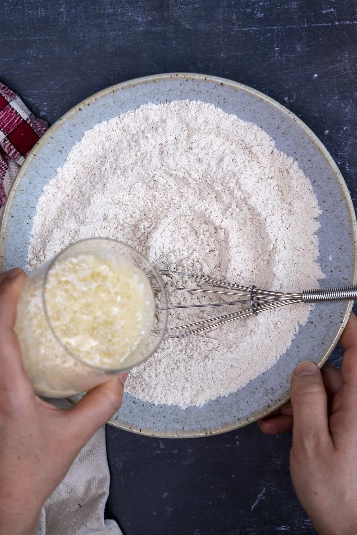 A hand pouring vegan buttermilk mixture into flour mixture in a blueish bowl and a hand whisk inside it.