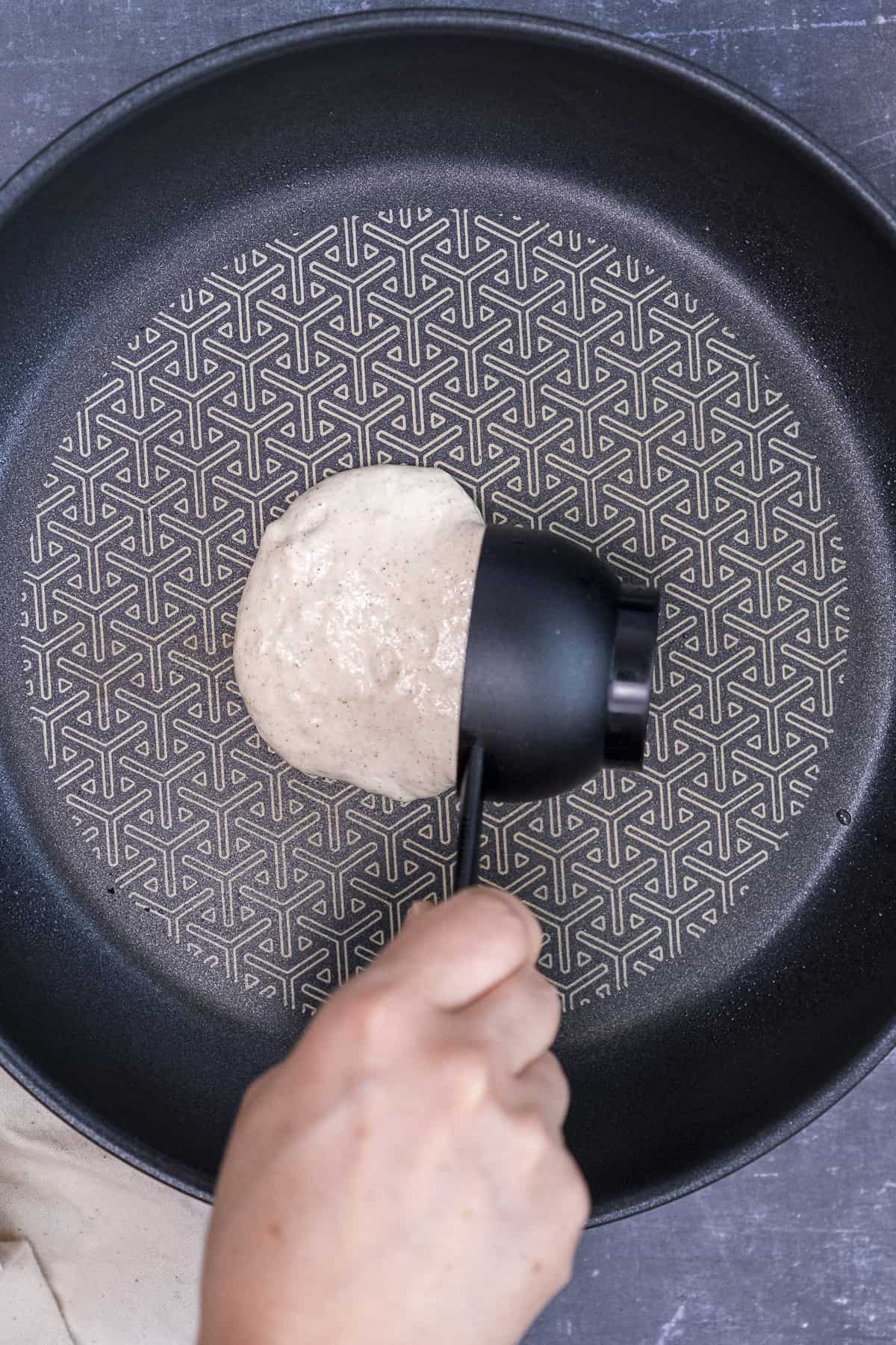 A hand pouring pancake batter from a measuring cup into a non-stick pan.